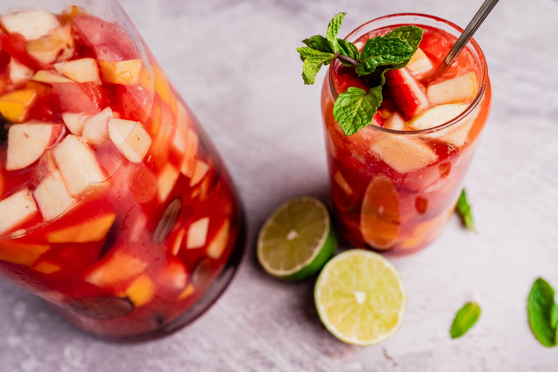 A glass and pitcher of refreshing tizana, brimming with mixed fruits, mint garnish, and a metal straw, sit beside halved limes on a textured surface.