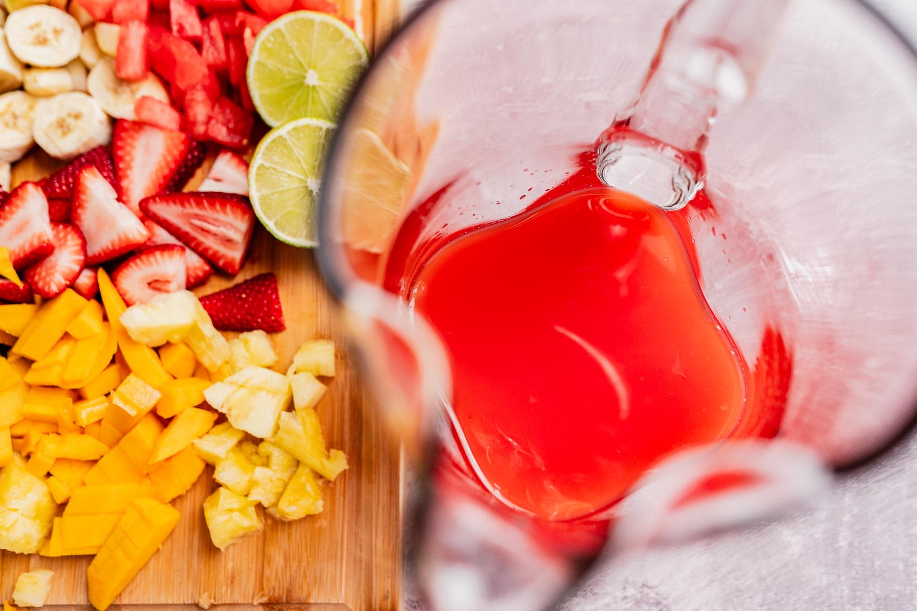 A glass pitcher brimming with tizana sits beside a wooden board adorned with sliced strawberries, bananas, mangoes, pineapple, and lime.