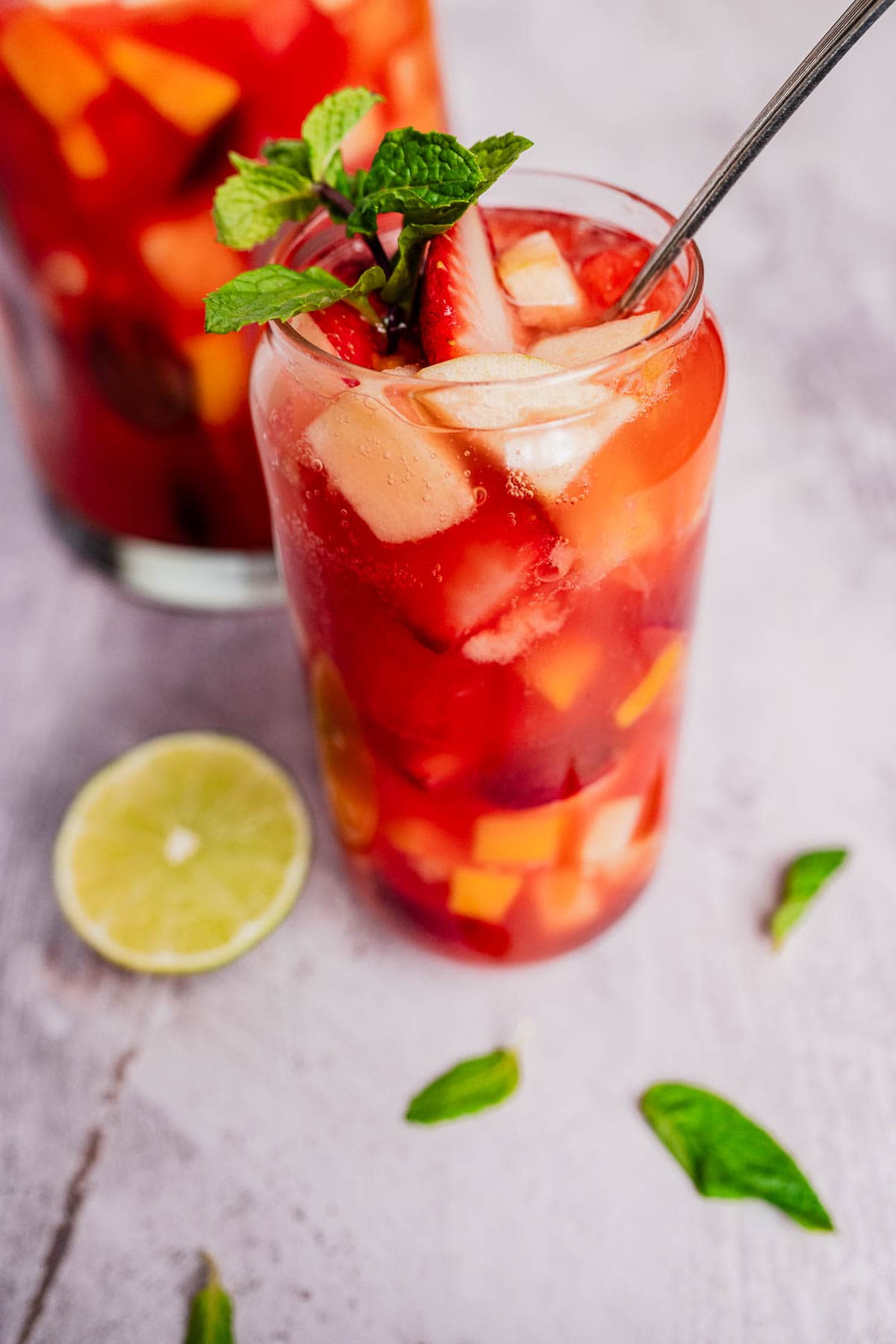 A glass filled with a vibrant red tizana brims with ice cubes, strawberry slices, and mint leaves. Nearby, a slice of lime and sprigs of mint are artfully scattered on the table.