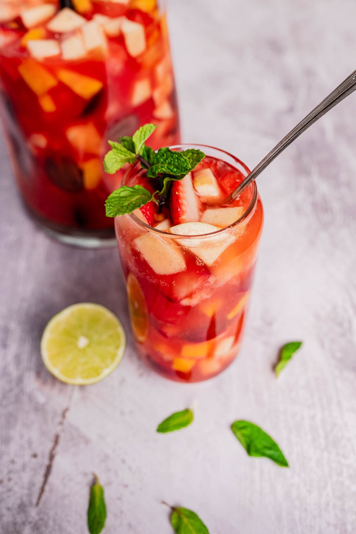 A glass of fruit-filled red sangria with a spoon, reminiscent of a vibrant tizana, garnished with mint leaves. A lime slice and mint leaves are scattered on a gray surface.