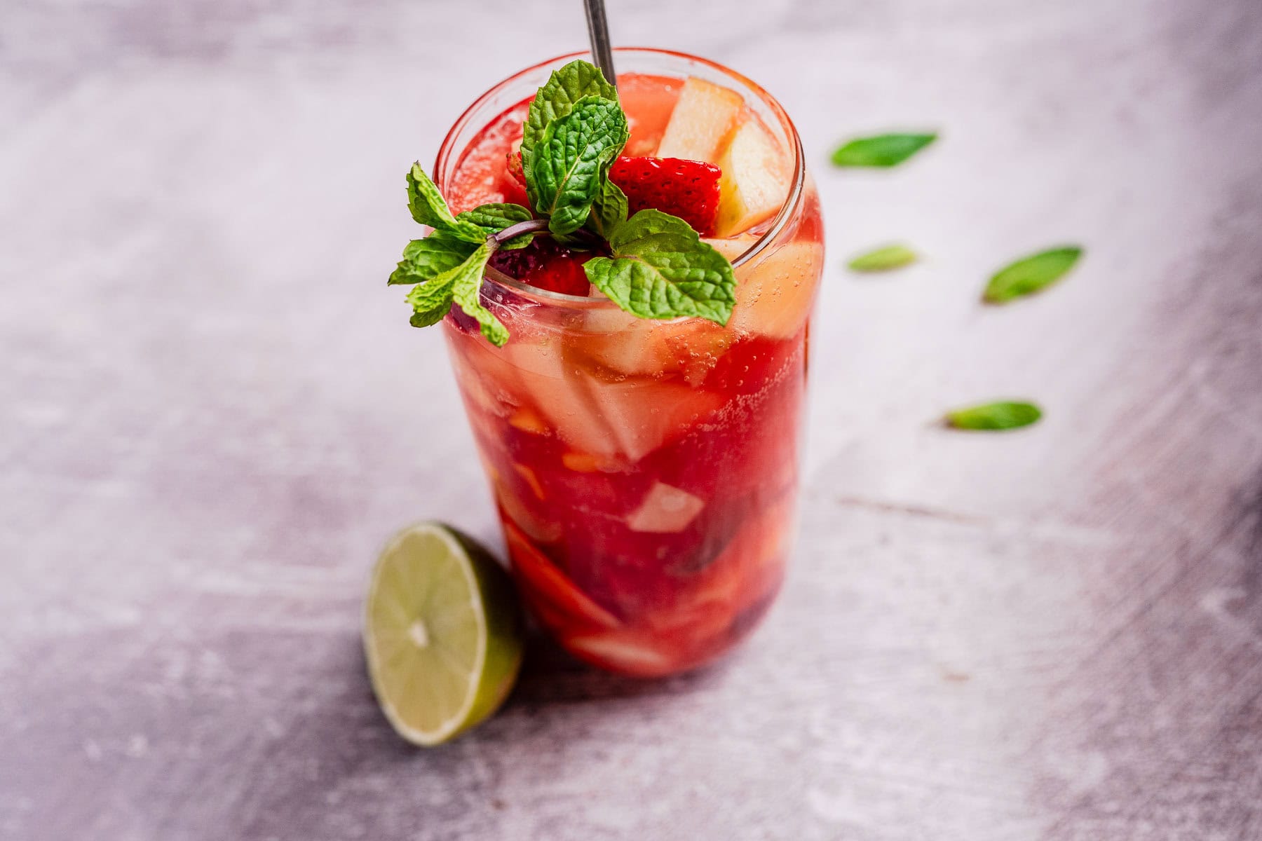 A glass of tizana, fruit-infused and chilled with ice, is elegantly garnished with mint and a strawberry, complemented by a lime half on the side, all resting on a textured surface.