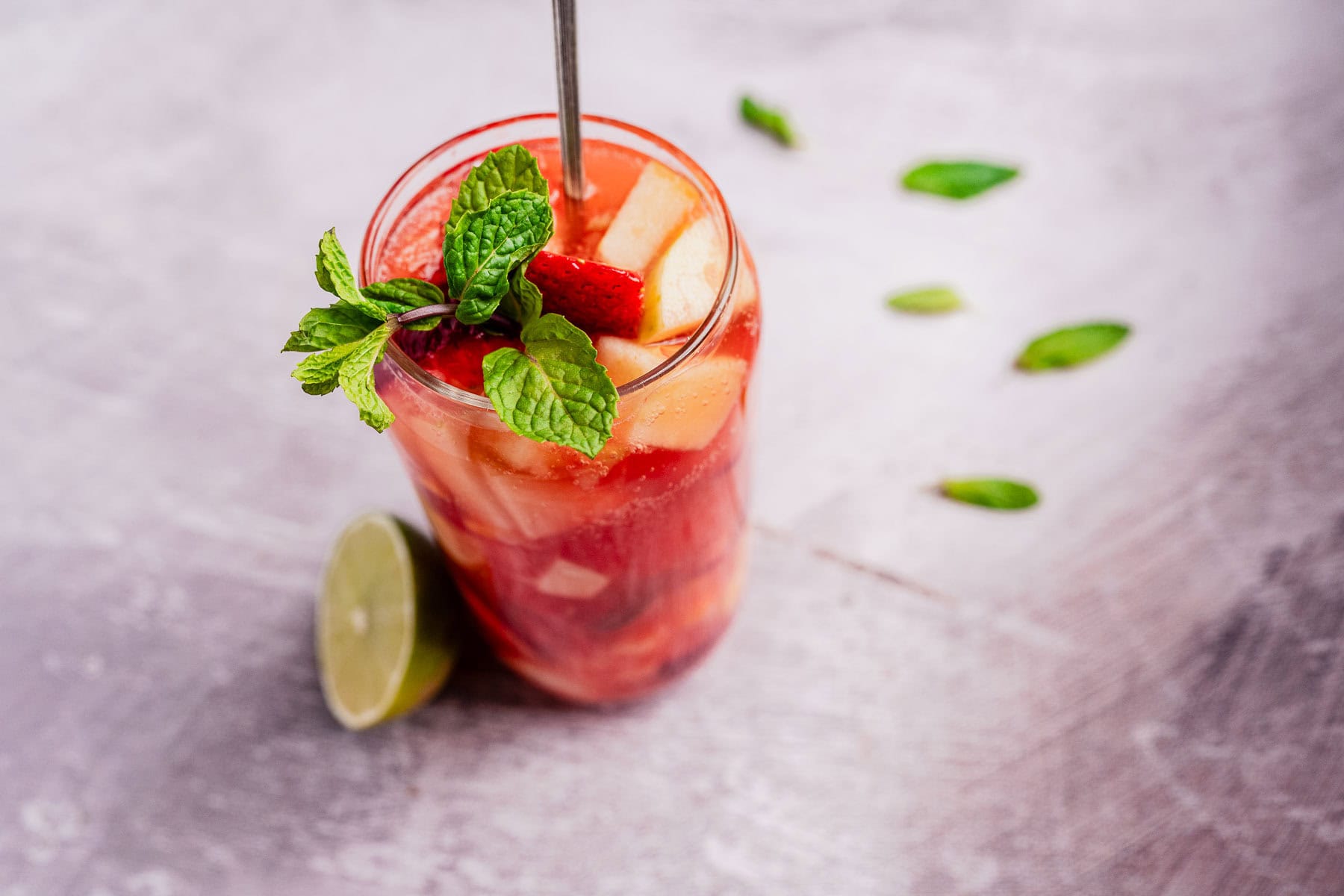 A tall glass brimming with a tizana, featuring a pink drink adorned with ice cubes, strawberry slices, and mint leaves. A metal straw stands ready in the refreshing mix. Beside the glass, a lime half and scattered mint leaves hint at more zesty flavors to come.