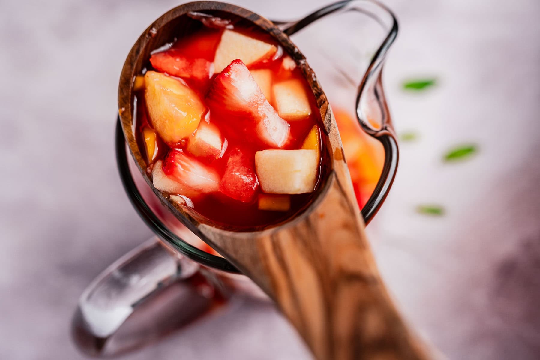 A wooden ladle brimming with diced fruit like strawberries, pineapple, and melon hints at the refreshing tizana within the glass pitcher backdrop.