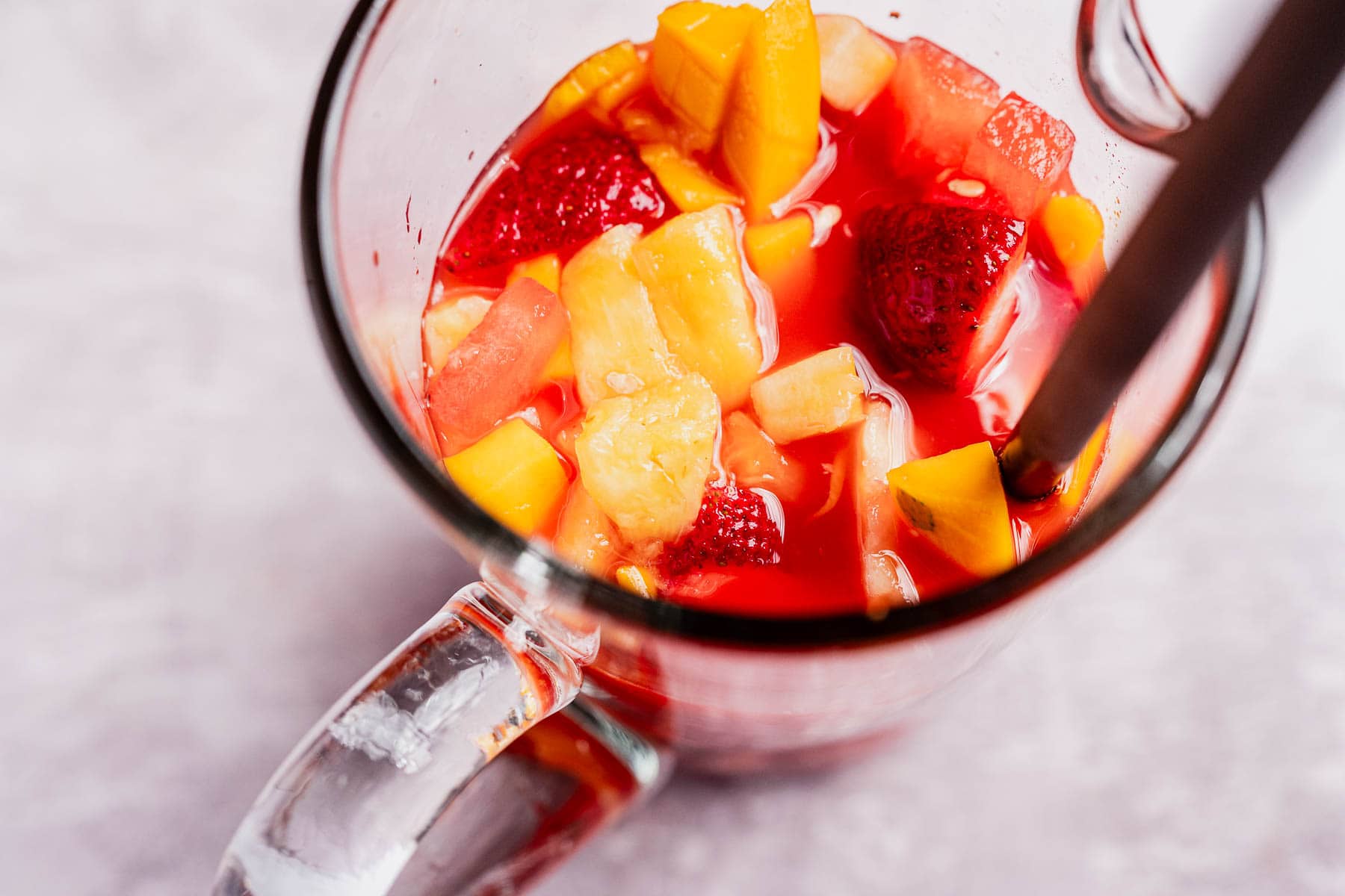 A glass mug brimming with a vibrant tizana, showcasing ice cubes and chunks of pineapple, mango, and strawberries, complete with a straw.
