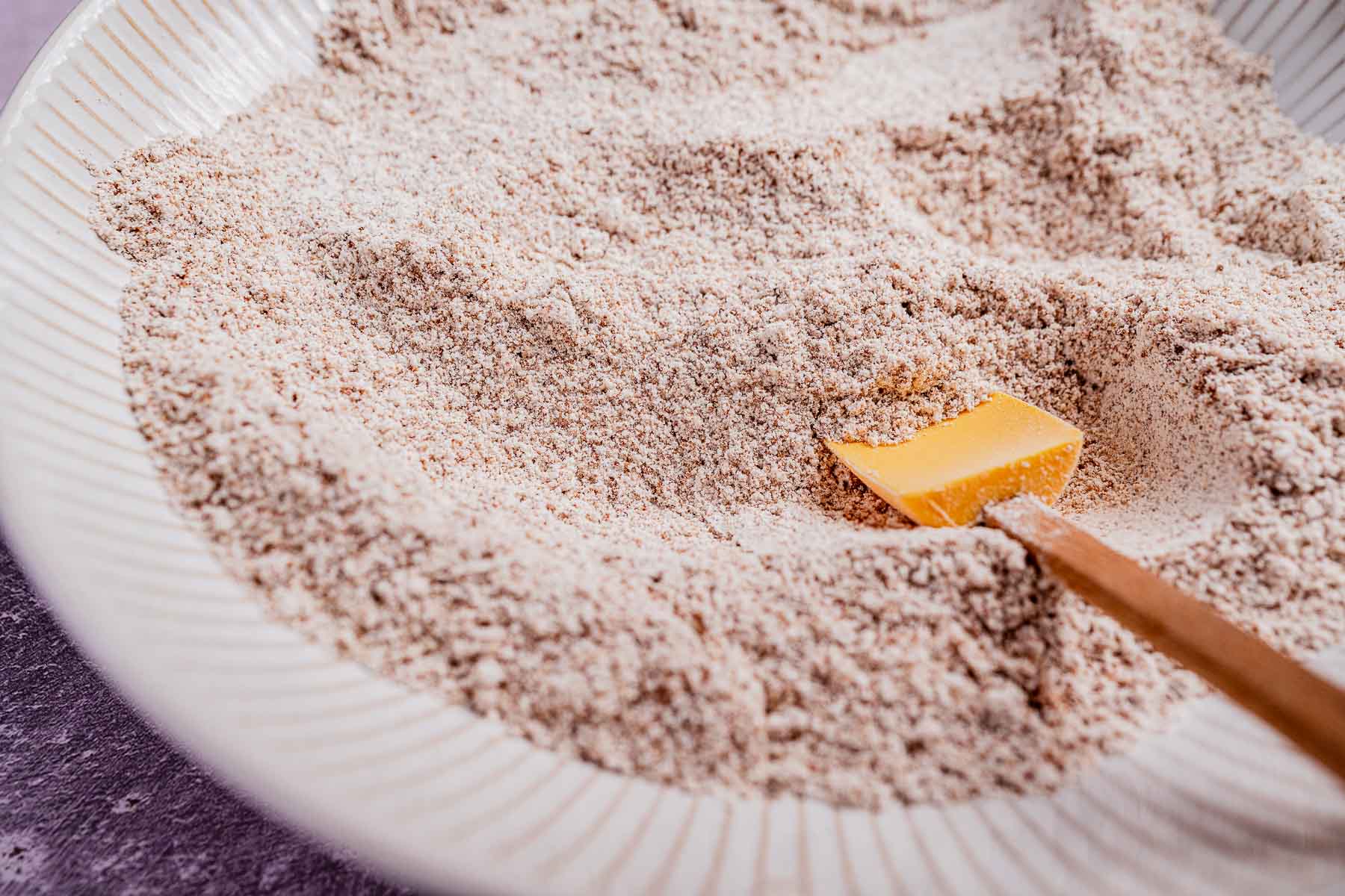 A bowl filled with grainy oat flour, with an orange spatula partially buried in it. The bowl is placed on a textured purple surface, ready to be transformed into delicious chocolate chip cookies.