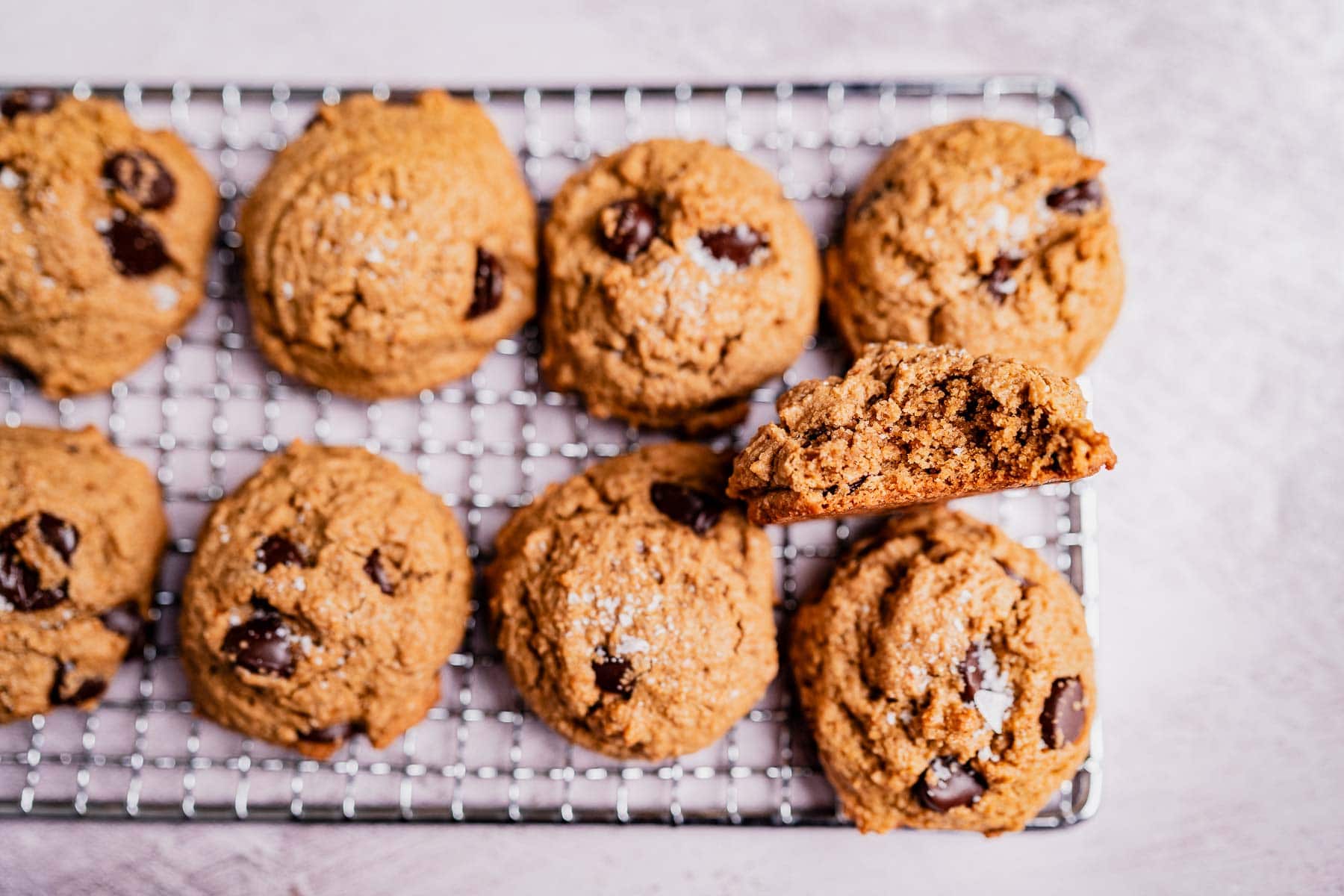 A cooling rack with oat flour chocolate chip cookies, one of which is broken in half to reveal its gooey inside.