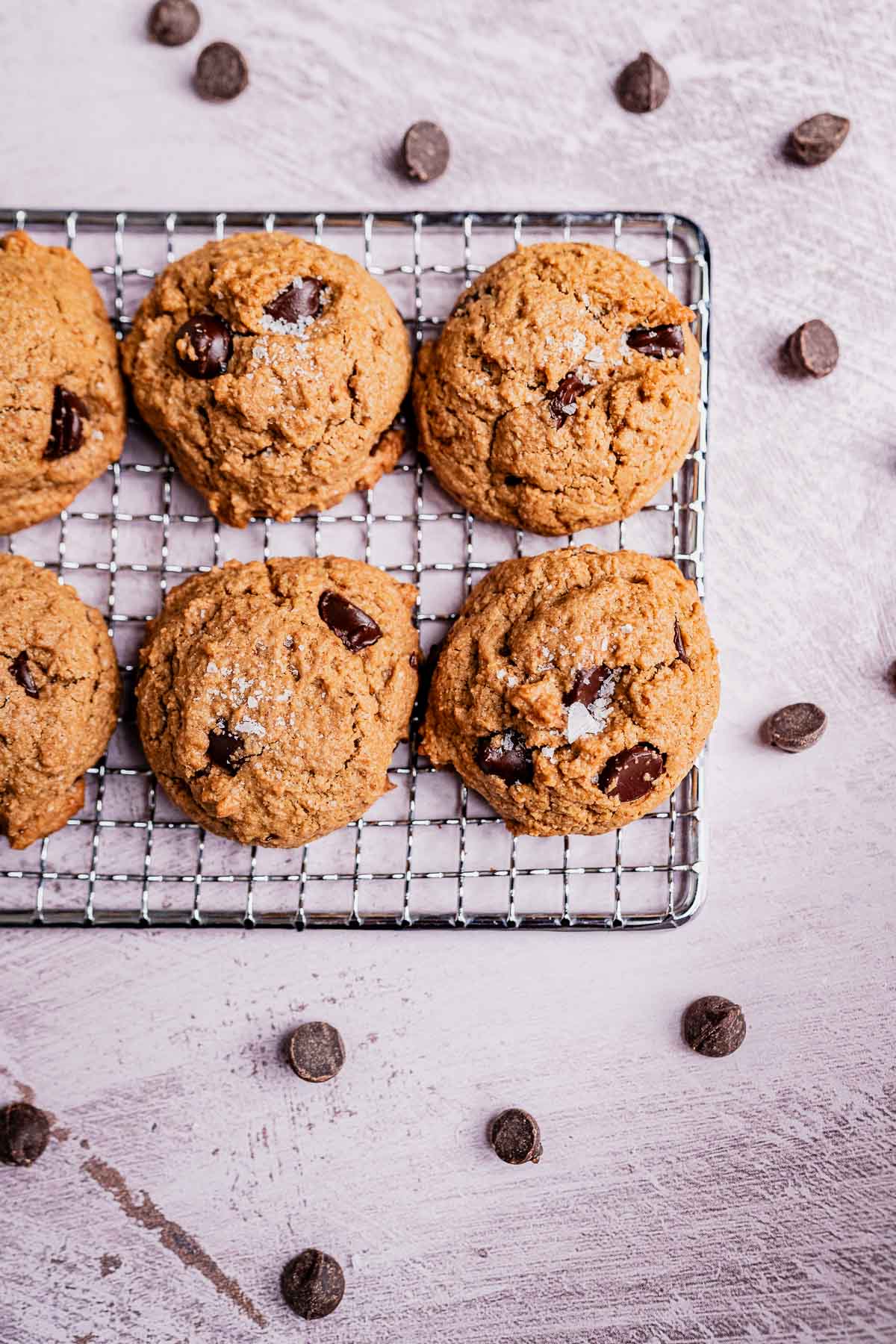 Six oat flour chocolate chip cookies rest on a cooling rack, surrounded by scattered chocolate chips on a light surface.