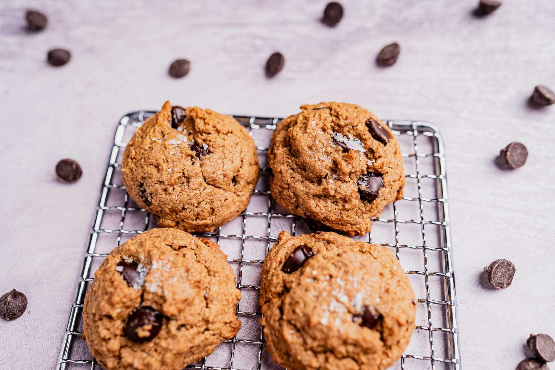 Four oat flour chocolate chip cookies rest on a cooling rack, surrounded by scattered chocolate chips on a light surface.