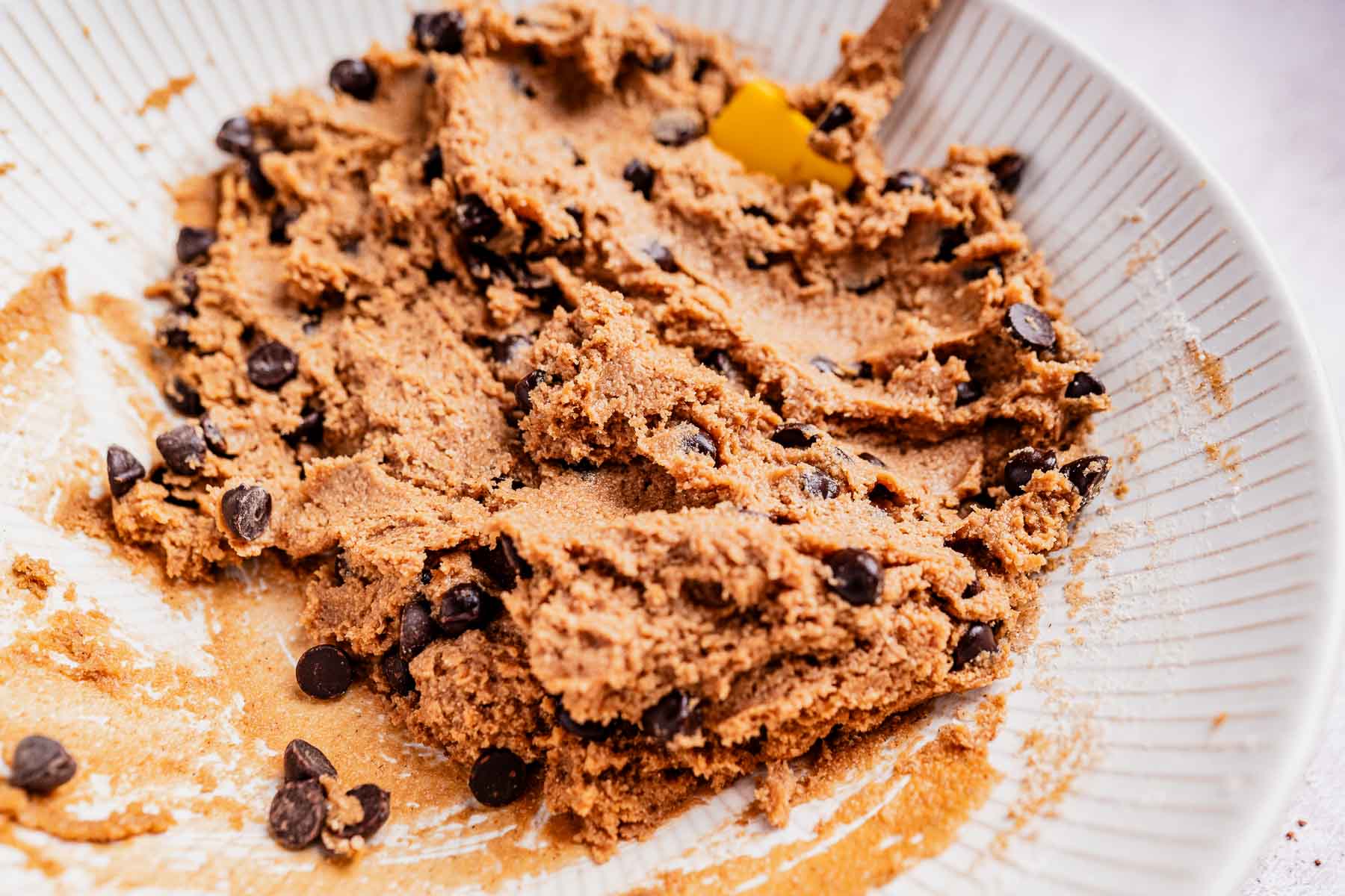 A bowl of raw oat flour chocolate chip cookie dough with a yellow spatula.