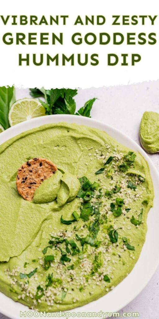 A bowl of green goddess hummus dip garnished with chopped herbs and seeds, with a round cracker dipped in, on a white surface alongside a wedge of lime and fresh basil leaves.