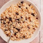 A plate of Arroz con Coco—cooked rice blended with raisins—is served with a spoon on a light-colored surface.