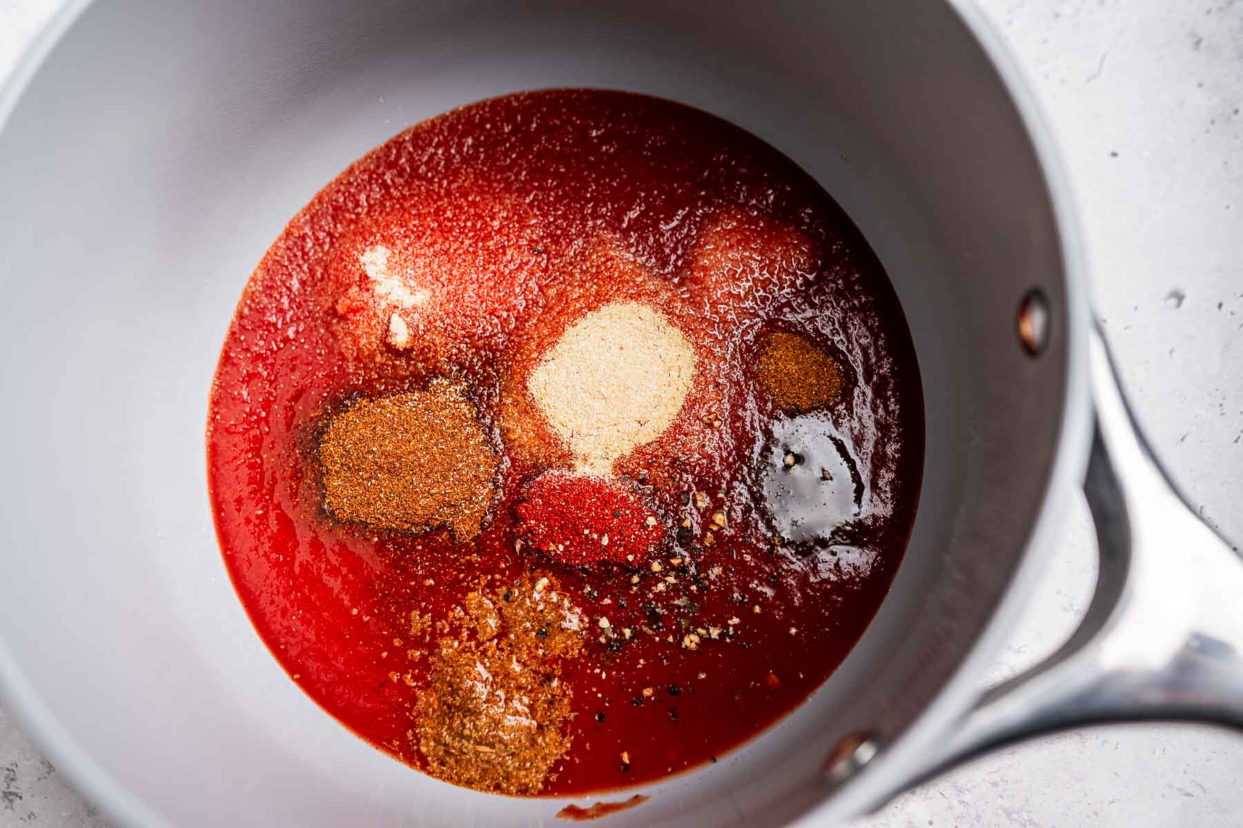 A saucepan filled with a red liquid and various spices, including pepper, paprika, and garlic powder, ready to be mixed into a zesty taco sauce.