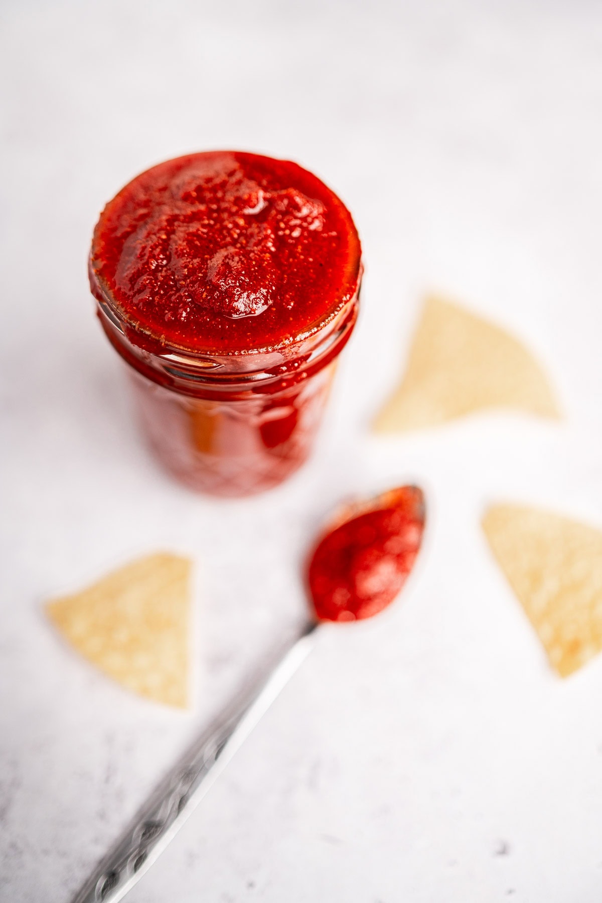 A jar of taco sauce with a spoonful beside it rests on a white surface, surrounded by three tortilla chips.