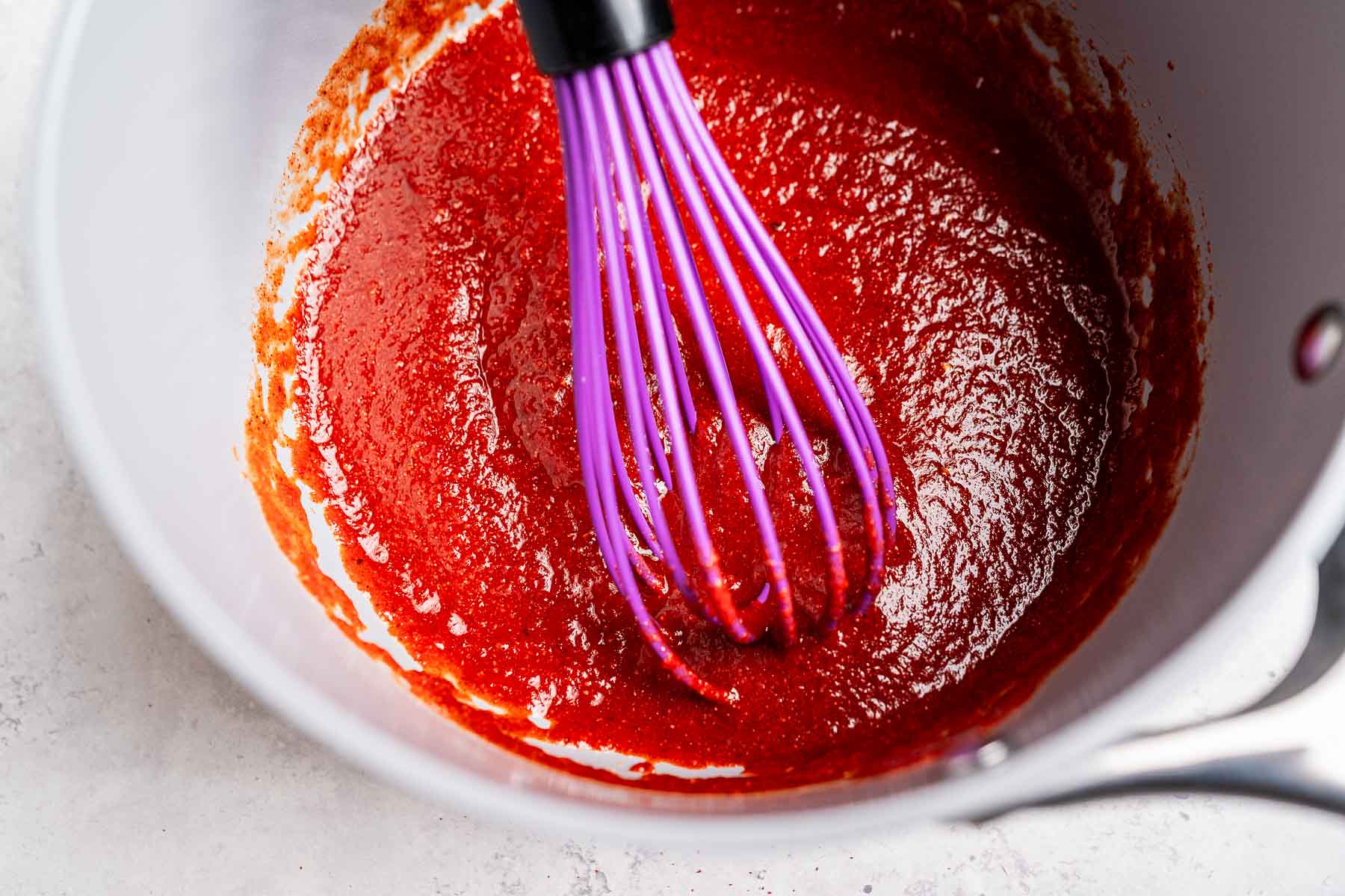 A purple whisk rhythmically blends a thick, red taco sauce in a metal pot on a light-colored surface.