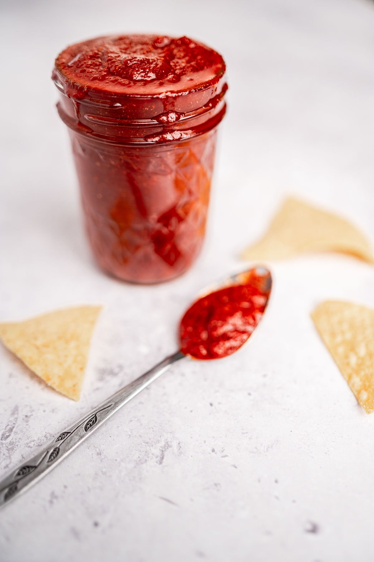 A jar of rich red taco sauce sits elegantly on a light surface, with a spoonful artfully placed beside it, surrounded by a tempting array of tortilla chips.