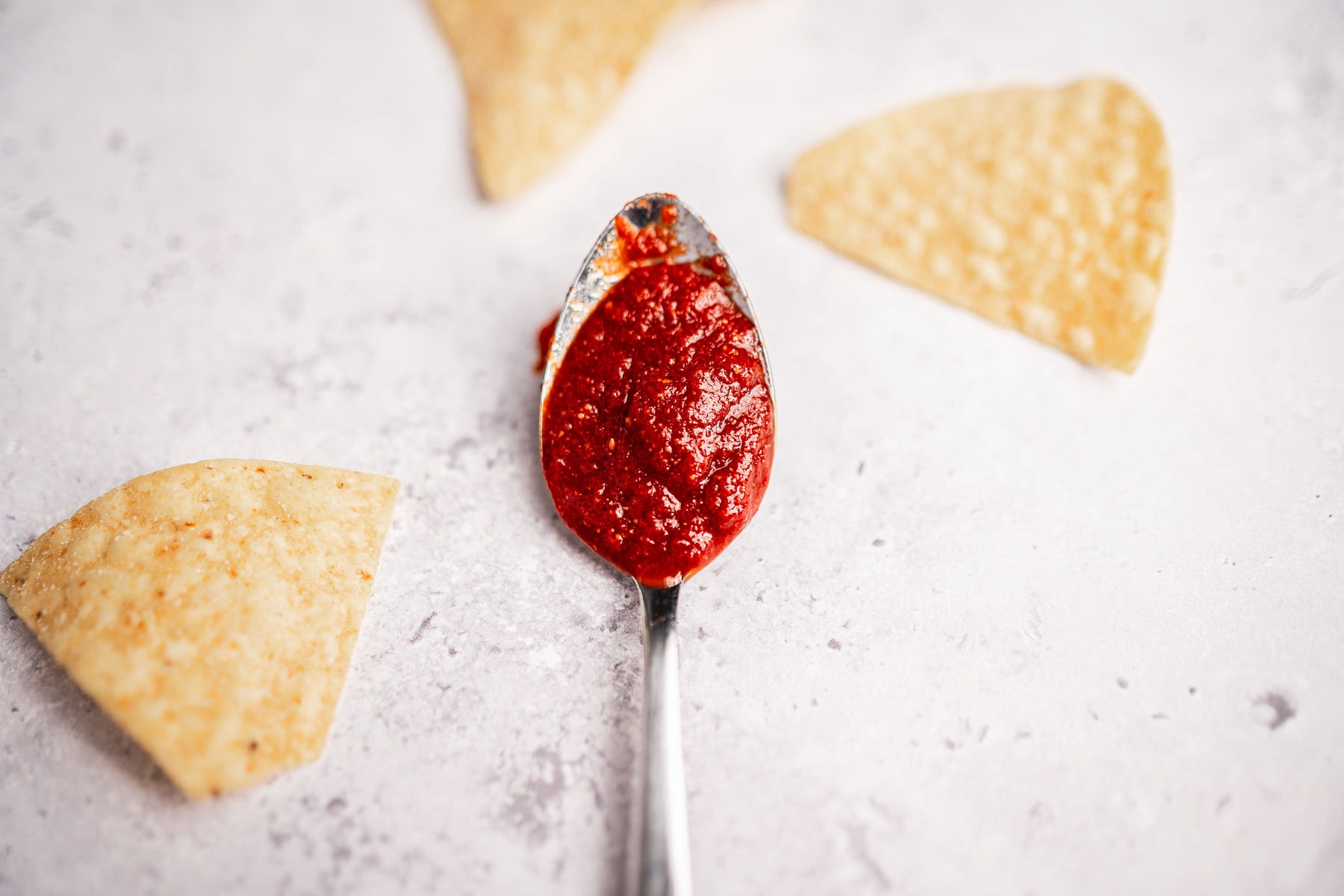 A spoonful of red salsa, reminiscent of rich taco sauce, is centered on a light surface, surrounded by three scattered tortilla chips.
