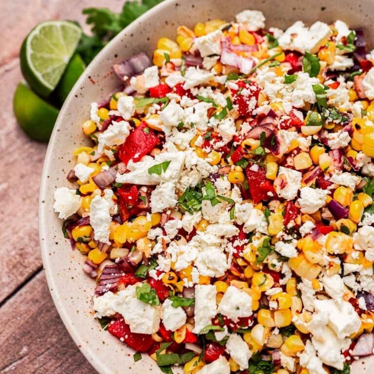 A bowl of charred corn salad with chopped red peppers, onions, cilantro, and crumbled white cheese, served with lime wedges on the side.