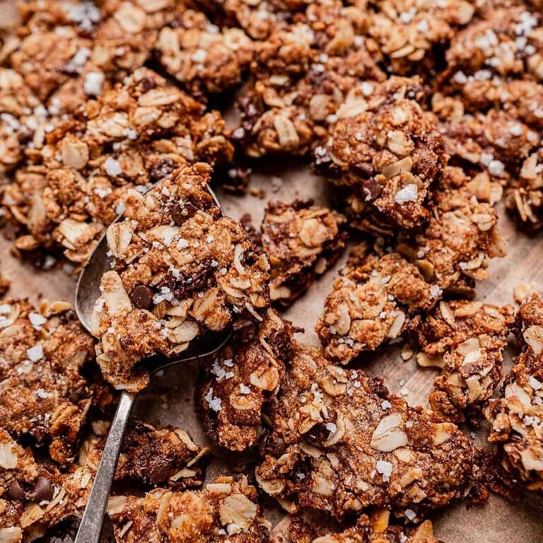 A close-up of chocolate chip cookie granola clusters on parchment paper, with a metal spoon lifting one piece.