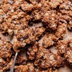 A close-up of chocolate chip cookie granola clusters on parchment paper, with a metal spoon holding one piece.