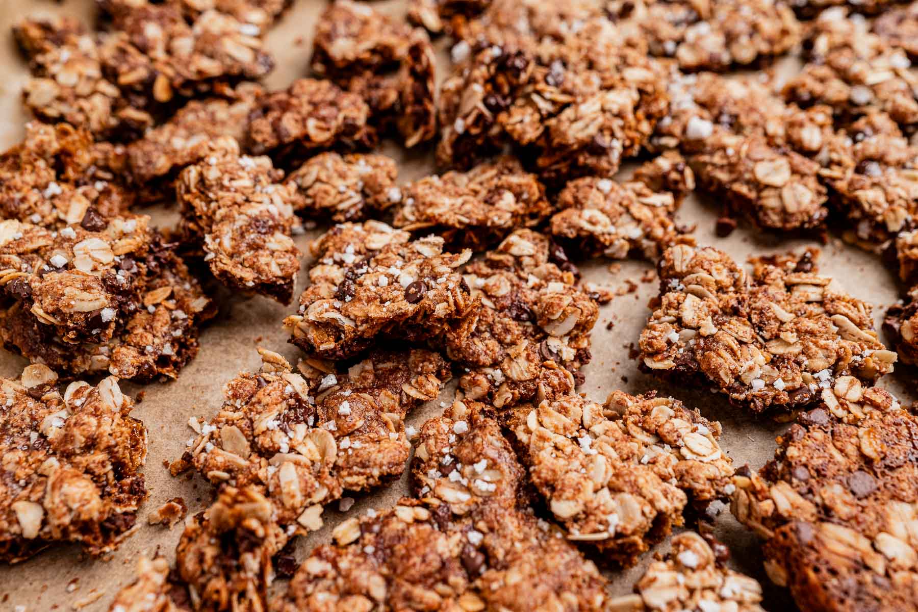 Close-up of chocolate chip cookie granola clusters on parchment paper, featuring a mix of oats, seeds, and chocolate chips with a golden-brown, crispy texture.