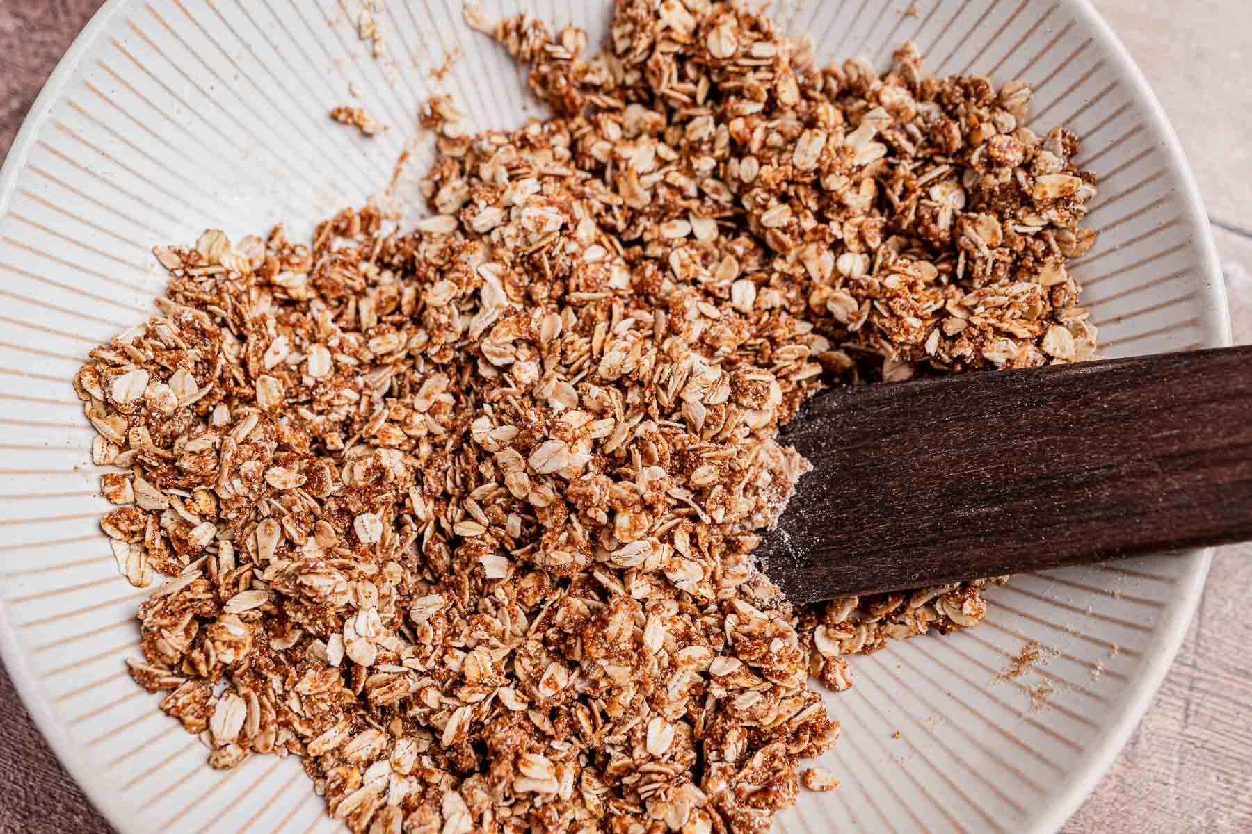 A bowl of chocolate chip cookie granola dry oat mixture is being stirred with a dark wooden spatula.