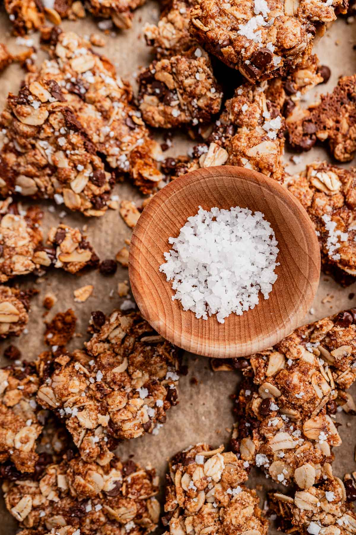 A wooden bowl filled with coarse sea salt sits on top of clusters of chocolate chip cookie granola and oat granola, all resting on a sheet of parchment paper.