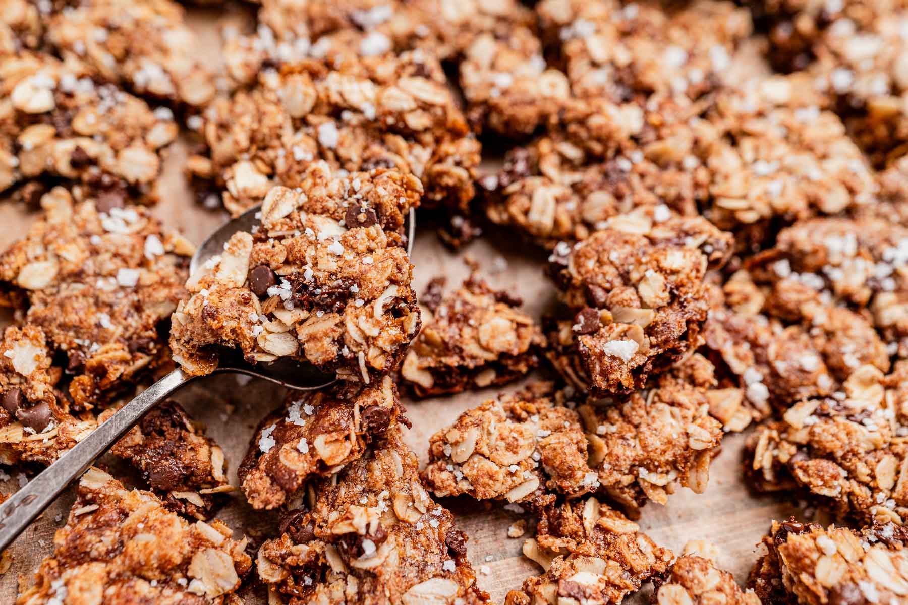 A close-up of several oatmeal chocolate chip cookie granola treats on a baking sheet, with a spoon holding one cookie in the foreground.
