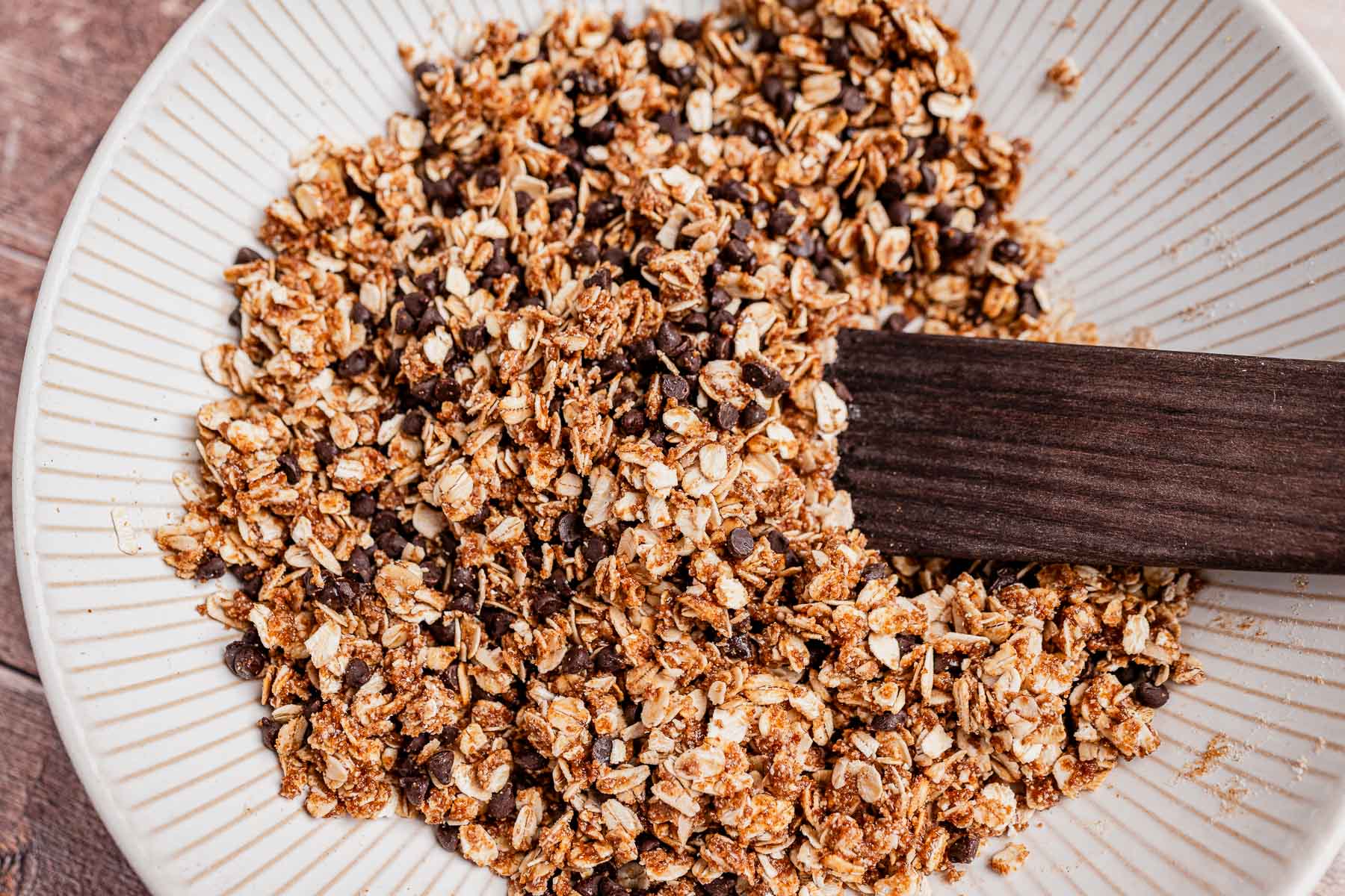 A close-up of a bowl containing chocolate chip cookie granola—a mixture of oats, chocolate chips, and spices—being partially stirred with a wooden spatula.
