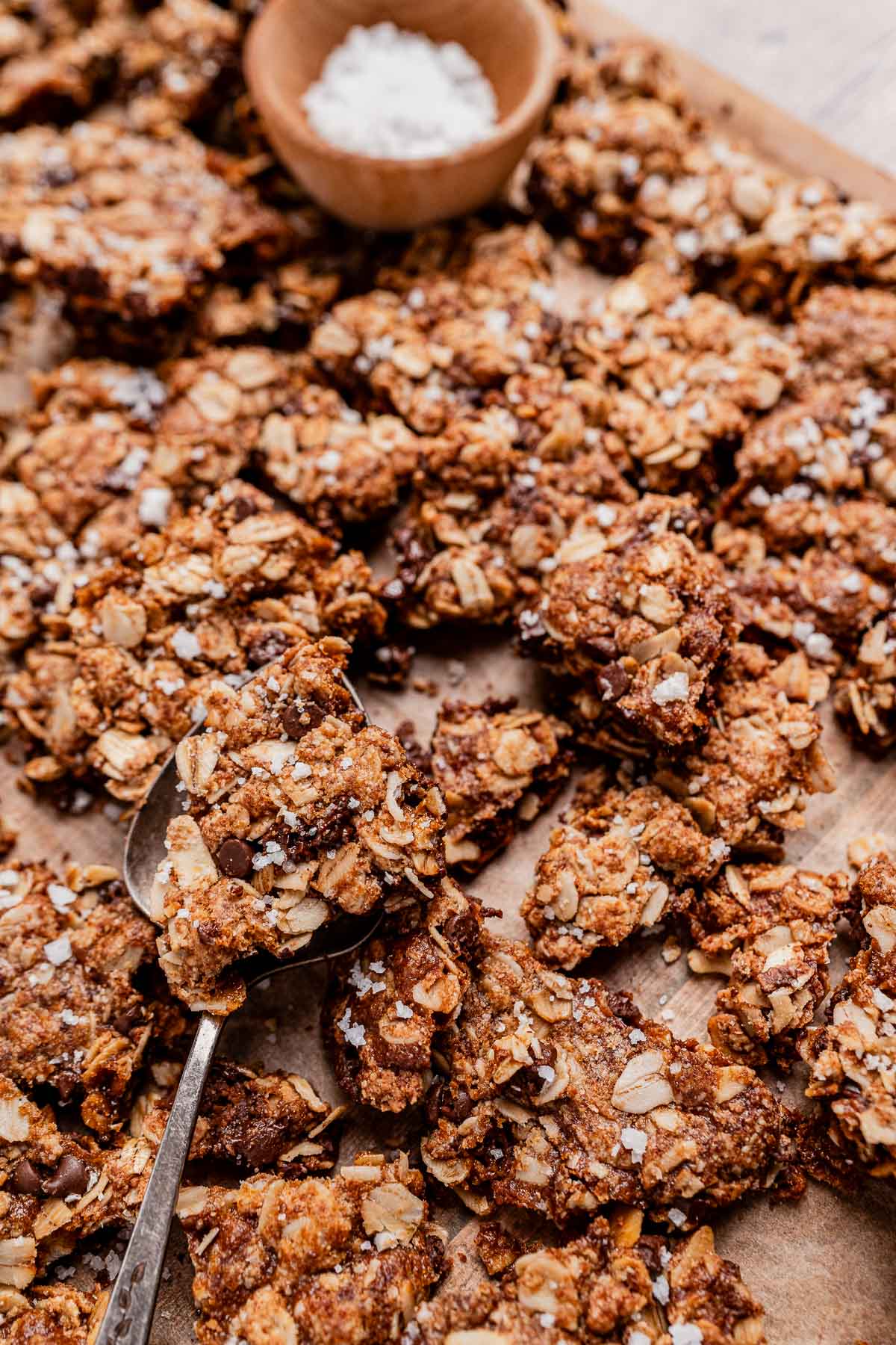 A close-up of oat and nut clusters on parchment paper, reminiscent of chocolate chip cookie granola, some sprinkled with coarse salt, with a spoon and a small wooden bowl of salt in the background.