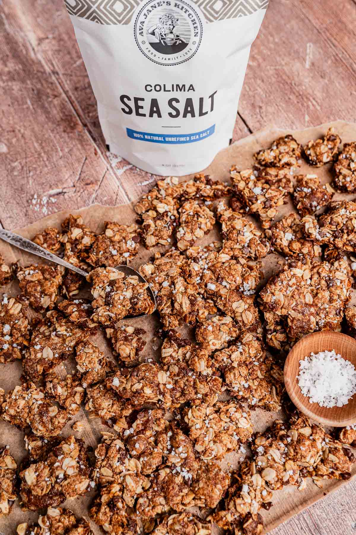 A batch of homemade chocolate chip cookie granola clusters on parchment paper with a spoon, a small wooden bowl of sea salt, and a bag of Colima sea salt in the background.