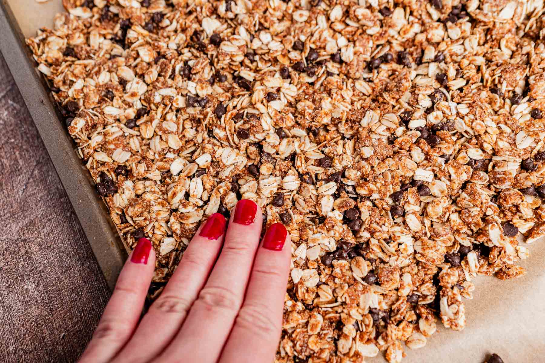 A hand with red nail polish presses down on a sheet of unbaked chocolate chip cookie granola mixture spread on parchment paper.