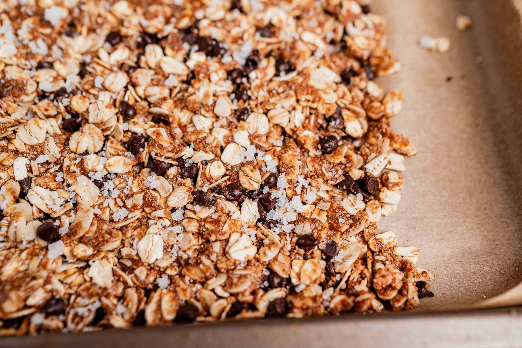 A close-up of a baking tray lined with parchment paper, filled with a chocolate chip cookie granola mixture of oats, chocolate chips, and coarse salt.