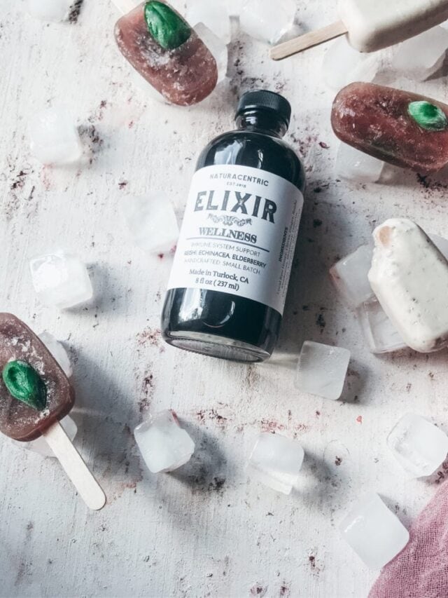A bottle labeled "Elixir Wellness Support" is surrounded by ice cubes and popsicles on a white surface.