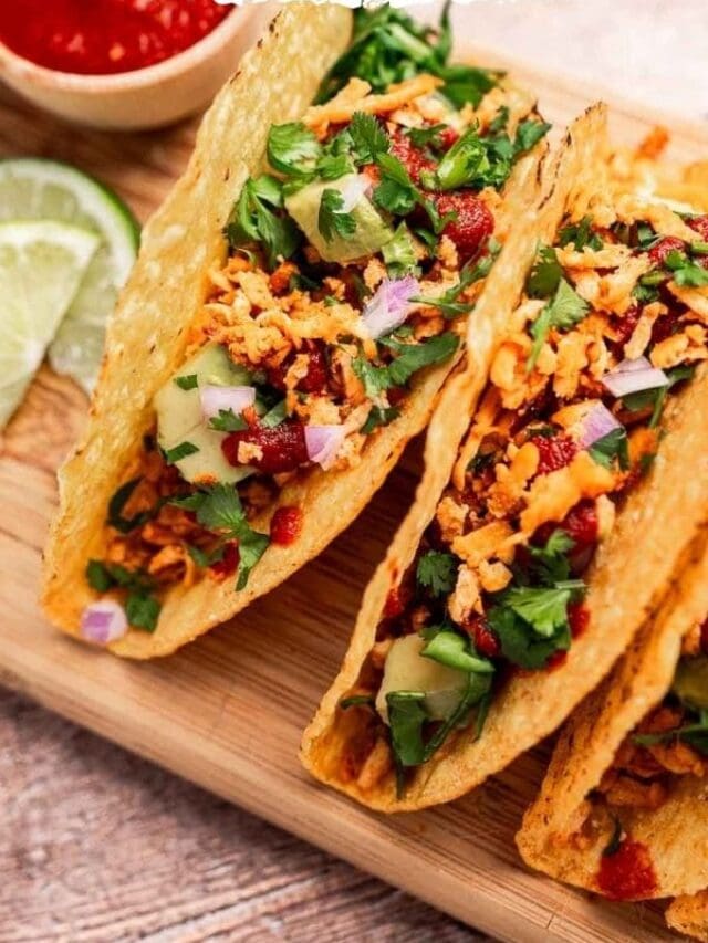 Three crispy tacos filled with seasoned meat, diced onions, cilantro, and sauce are served on a wooden board with lime wedges on the side.