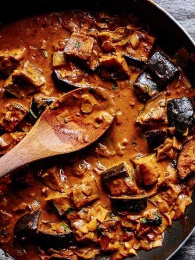 A skillet filled with a thick, brown vegetable stew, including chunks of eggplant and other vegetables, with a wooden spoon resting in the mixture.