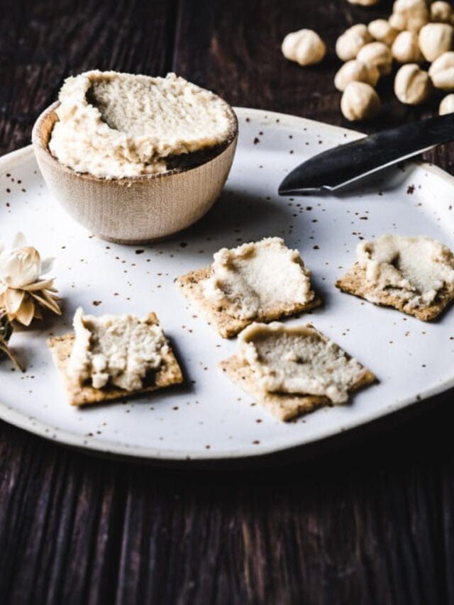 A bowl of nut spread with several crackers topped with the spread on a white plate. A knife and scattered nuts are in the background.