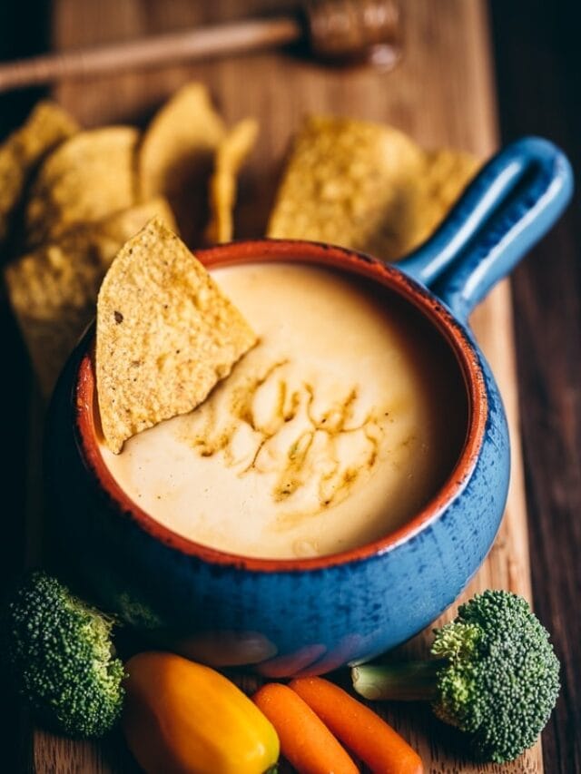 A blue bowl filled with cheese sauce, topped with a tortilla chip. Surrounding the bowl are tortilla chips, broccoli, carrots, and a small yellow bell pepper on a wooden surface.