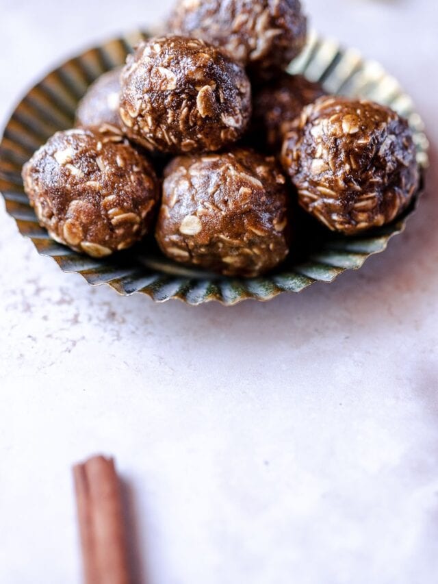 A small metal dish holds a stack of round, brown energy bites with visible oats. A cinnamon stick is placed on the marble surface nearby.