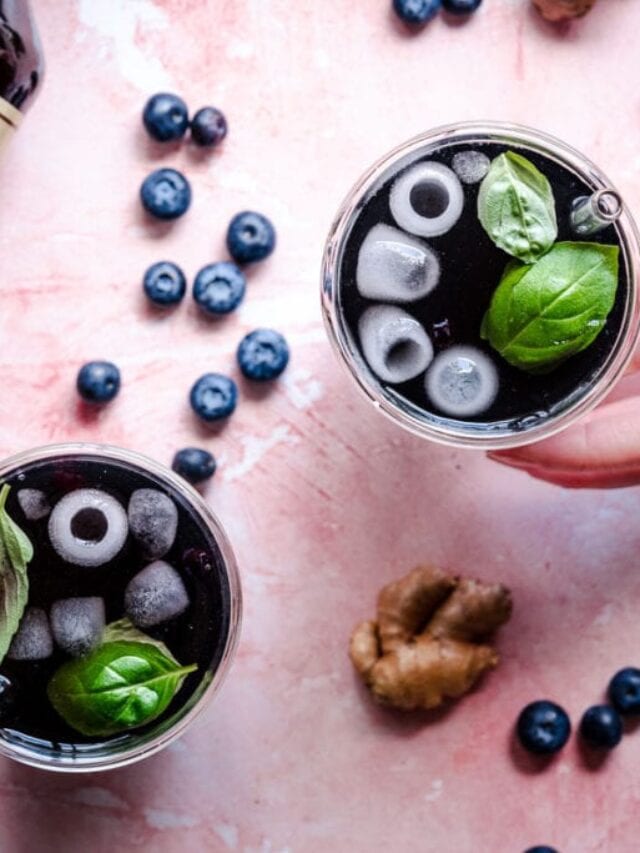 Two glasses of dark iced beverages with ice cubes and basil leaves, surrounded by scattered blueberries and ginger on a pink surface.