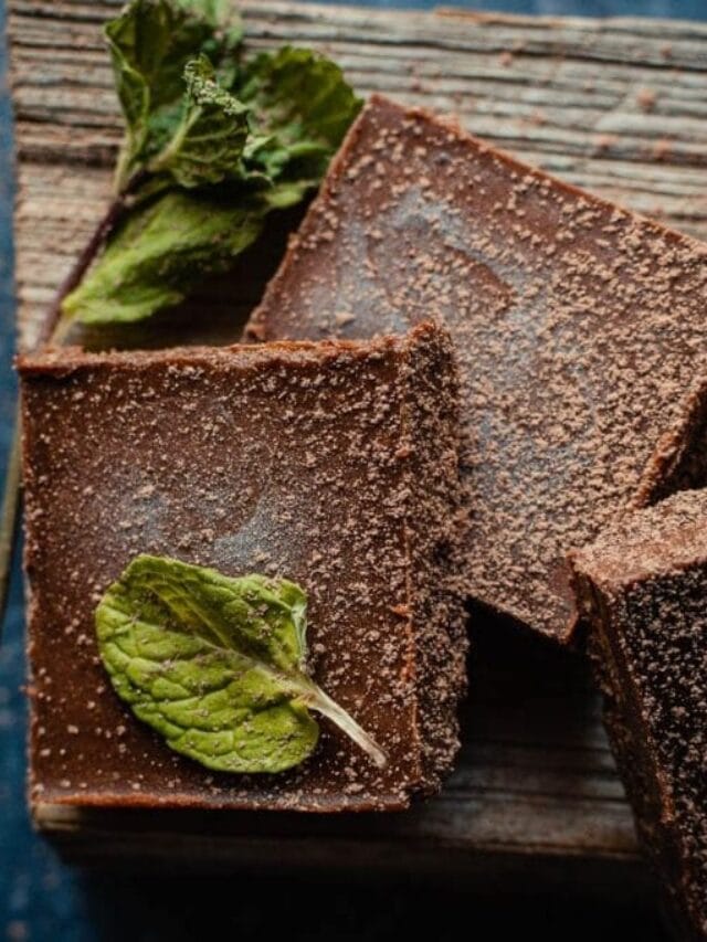 Two chocolate brownies with a dusting of cocoa powder, topped with mint leaves, placed on a wooden surface.