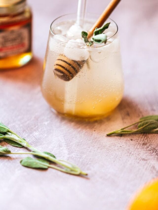 A glass of iced lemonade garnished with sage leaves and a honey dipper, next to fresh sage and a jar of honey on a table.