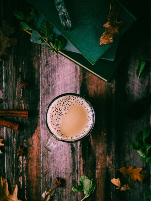 A glass mug filled with frothy coffee sits on a wooden table surrounded by scattered leaves, herbs, and two closed books.