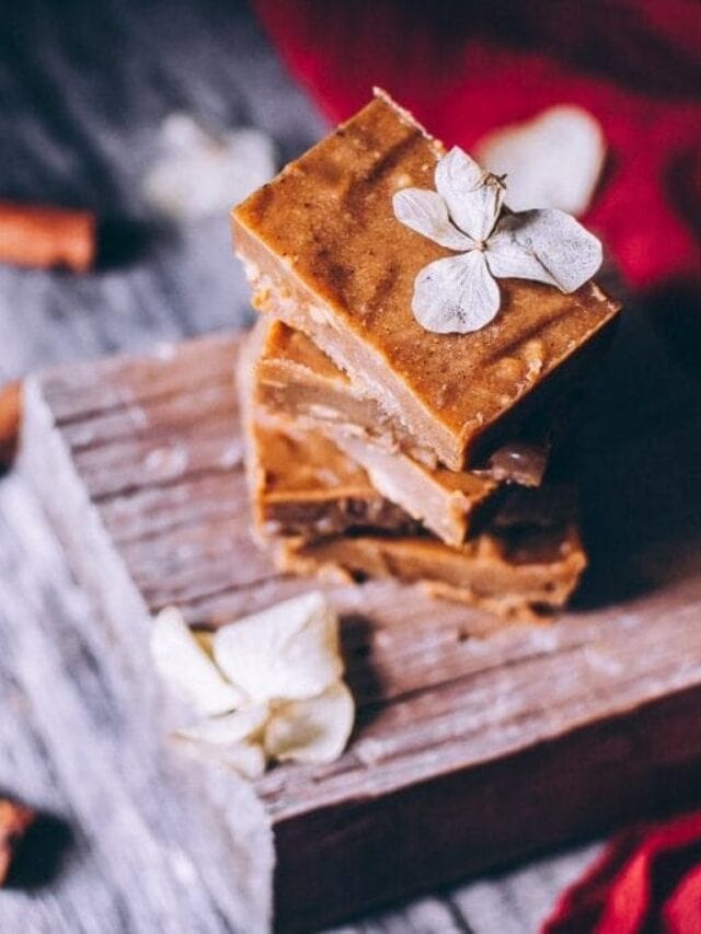 Three stacked pieces of brown fudge are placed on a wooden board, decorated with dried white flowers, with a red cloth and cinnamon sticks in the background.