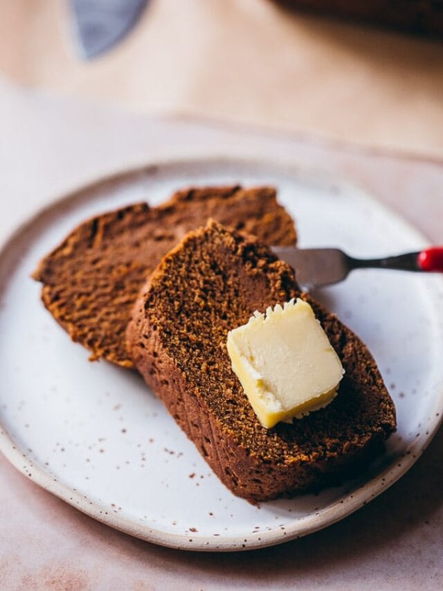 Two slices of brown bread on a white plate, topped with a pat of butter and a butter knife resting beside them.