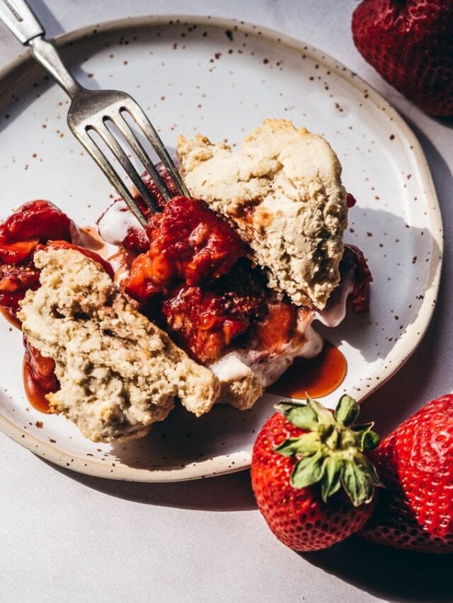 A fork rests on a plate with a strawberry shortcake topped with strawberries and syrup. Two fresh strawberries are beside the plate.