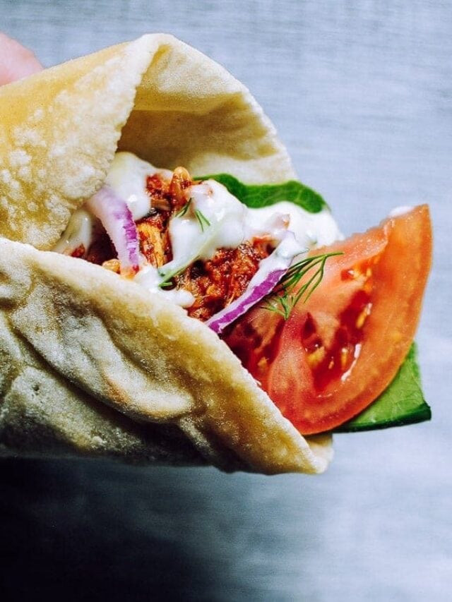 Close-up of a hand holding a pita wrap filled with shredded meat, sliced tomato, cucumber, red onion, greens, and a dollop of white sauce.