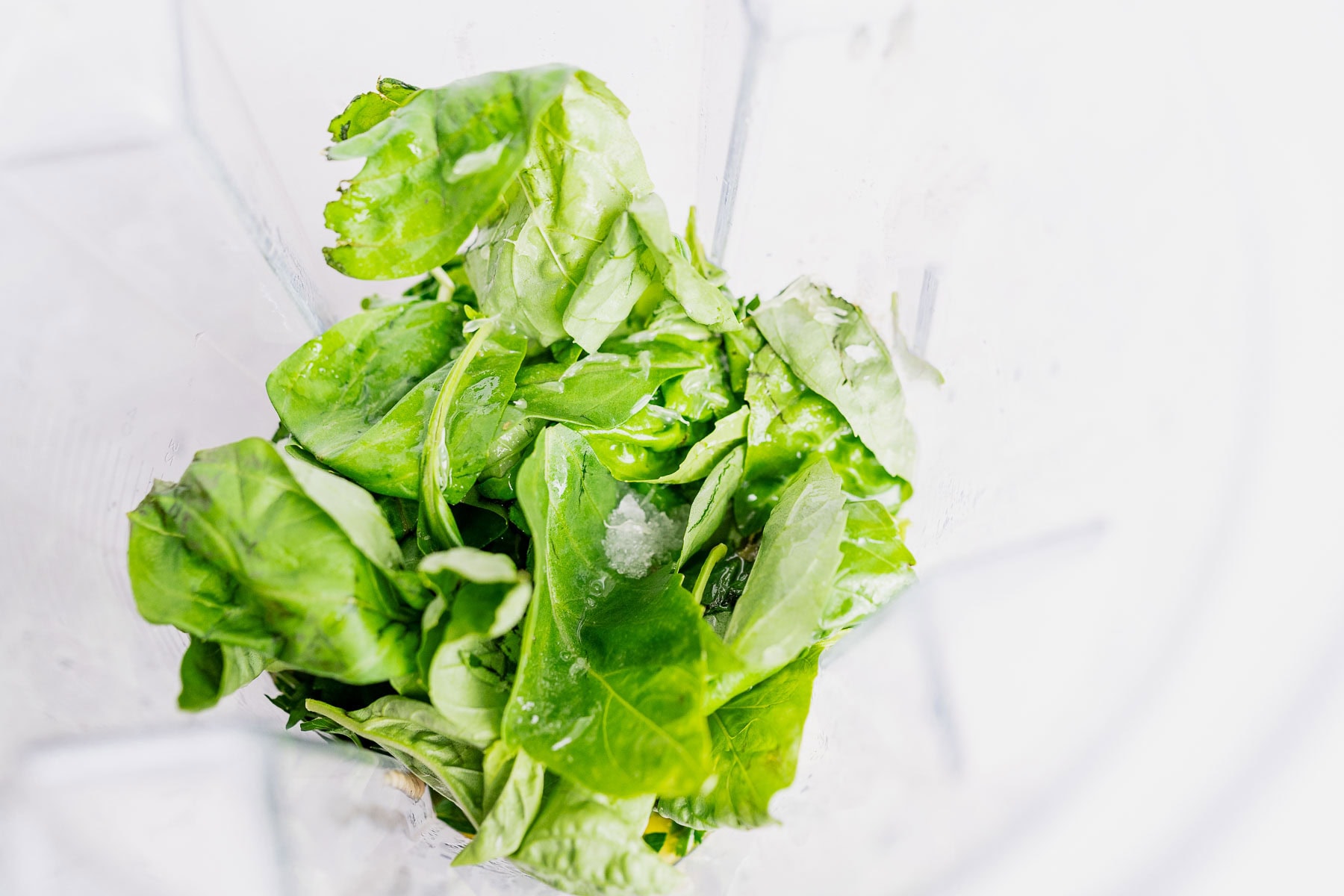 Fresh spinach leaves with a few ice cubes in a clear blender container, ready to whip up vibrant green goddess hummus, set against a white background.