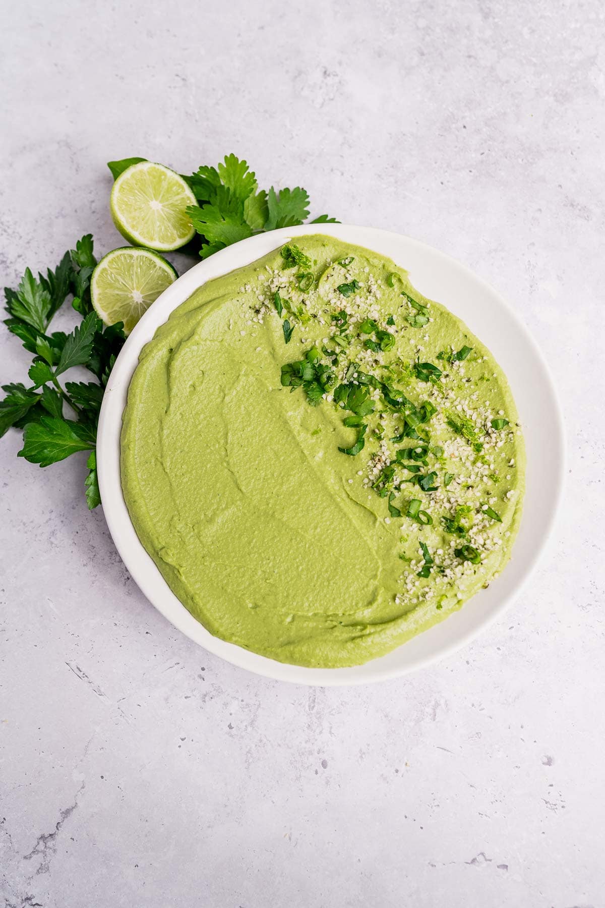 A bowl of green goddess hummus garnished with chopped herbs sits on a white surface, with parsley and two lime halves beside it.