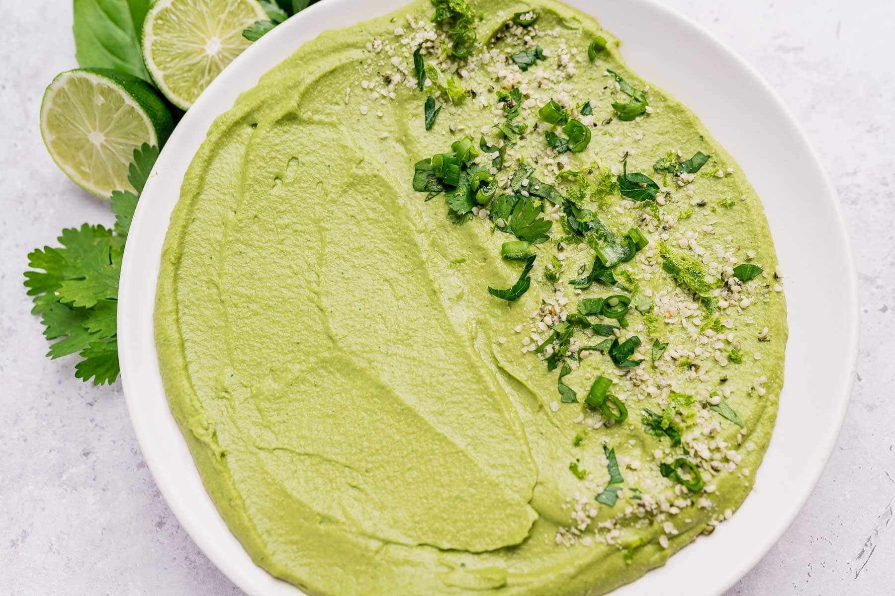 A white plate of green goddess hummus garnished with chopped herbs and seeds, with lime slices and cilantro on the side.