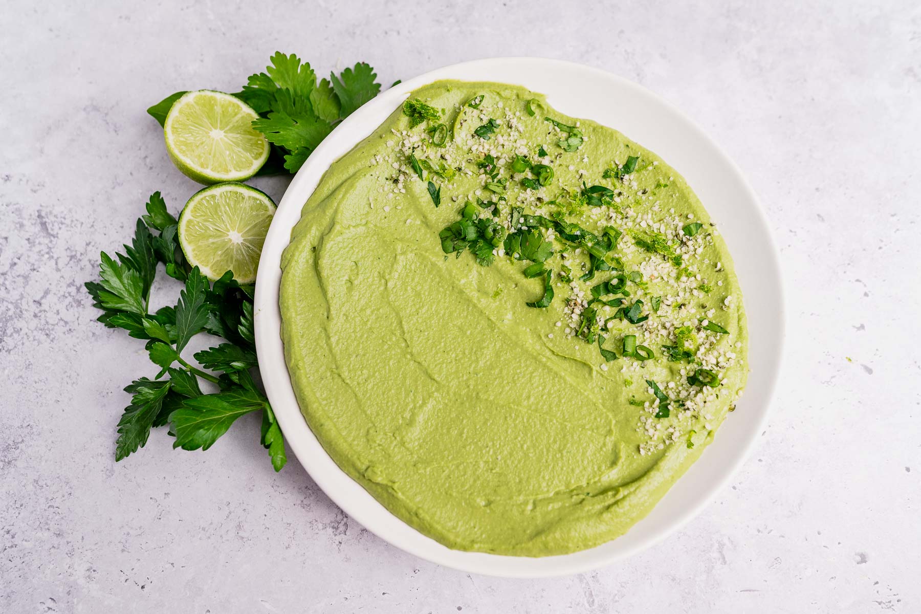 A white plate of green goddess hummus garnished with chopped herbs and seeds, with parsley and halved limes on the side, sits on a light textured surface.